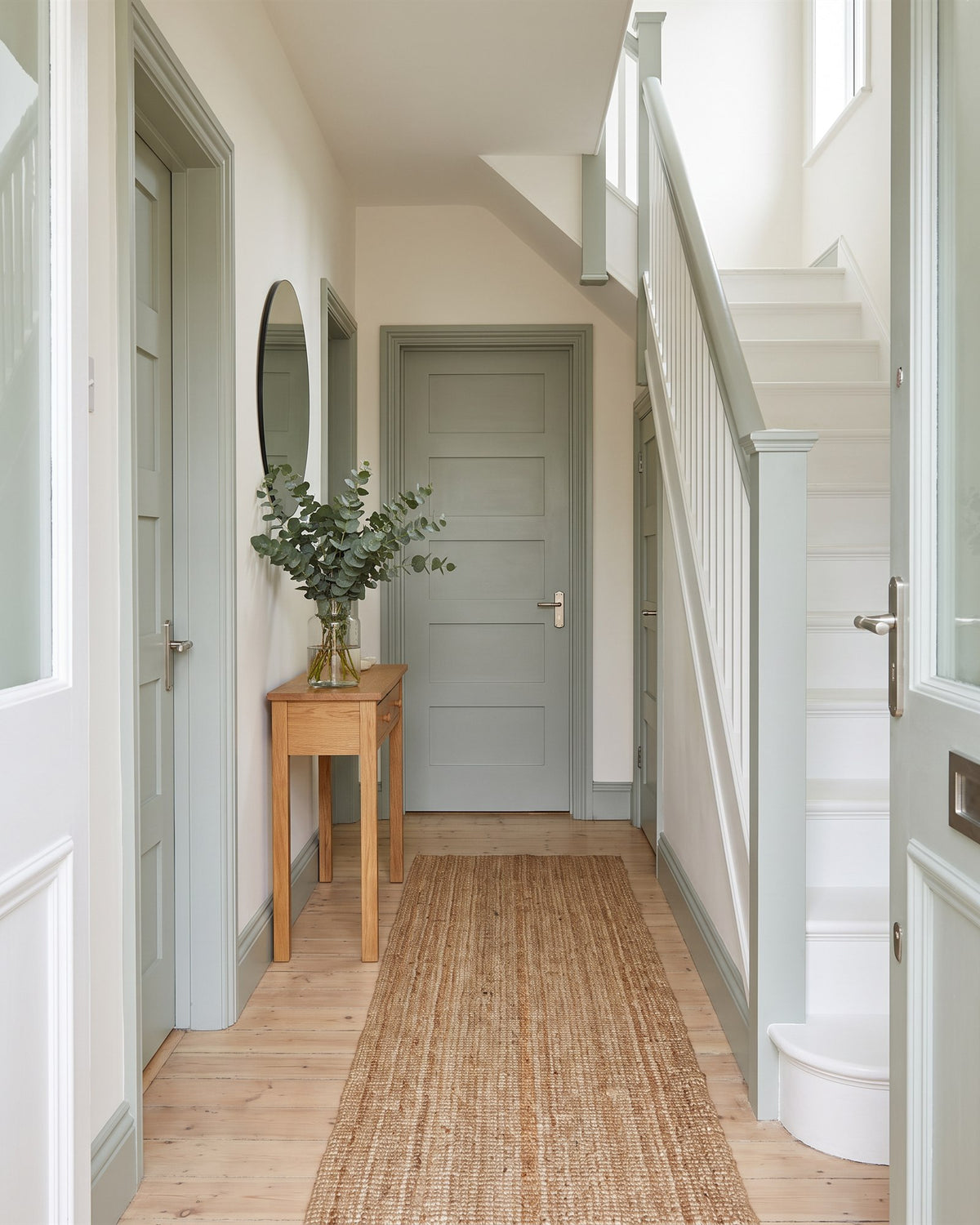 1920s hallway with warm white walls, pale oak boards, a jute runner, a white staircase with sage handrail and silvery sage doors with satin nickel handles