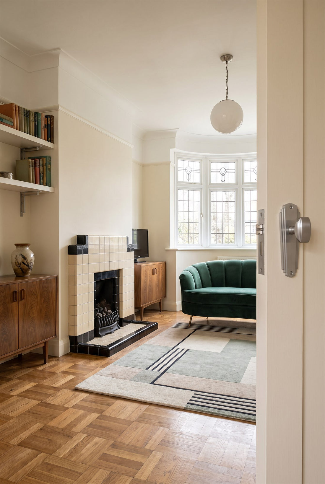 Restored 1930s lounge with a cream and black tiled fireplace, basket-weave parquet and green velvet sofa, satin chrome Art Deco shelf brackets in the alcove and a satin chrome Deco knob on plate on the door