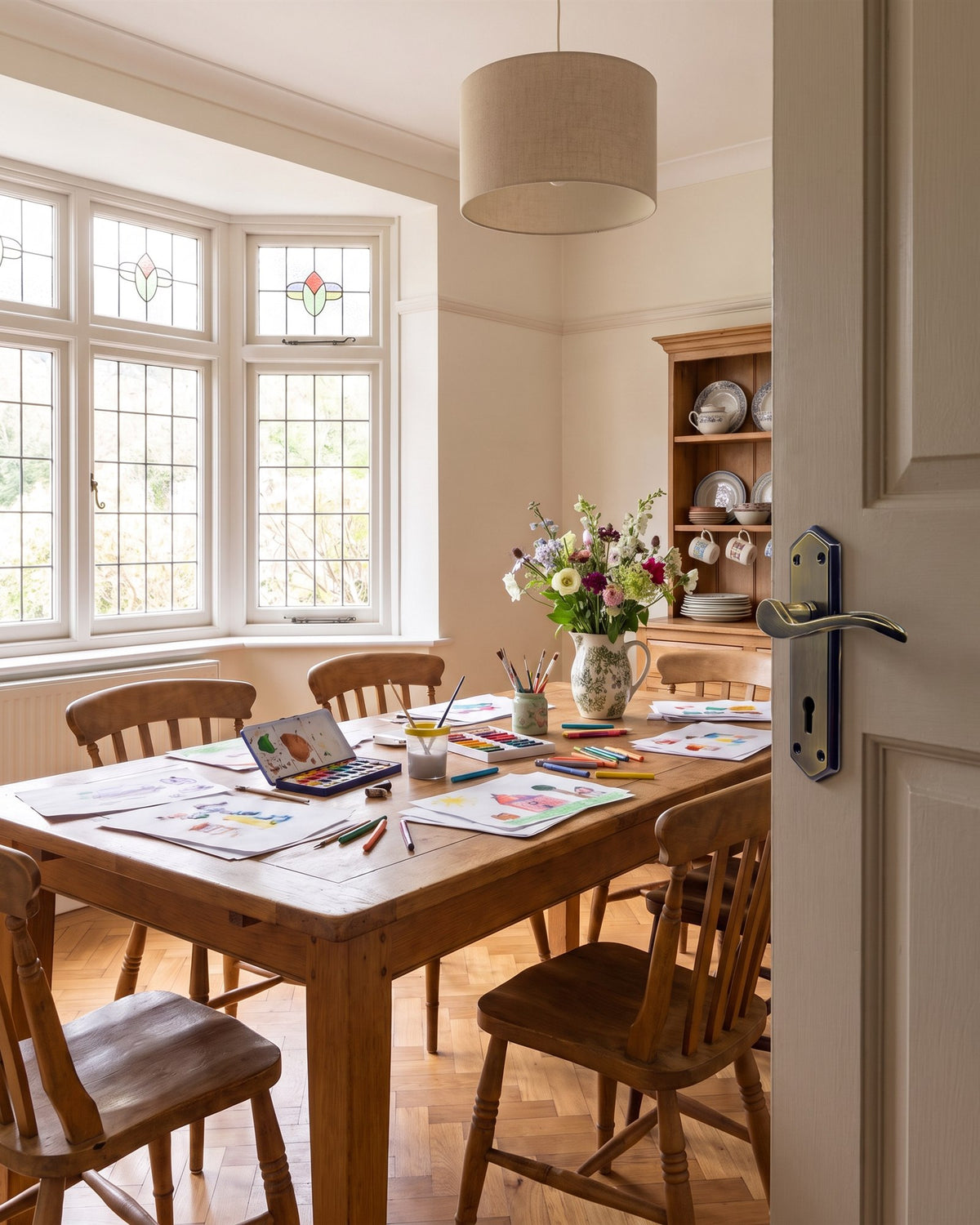 1930s dining room with a leaded bay window, pine table and dresser, seen past a greige door with an antique brass and bronze Paris lock handle