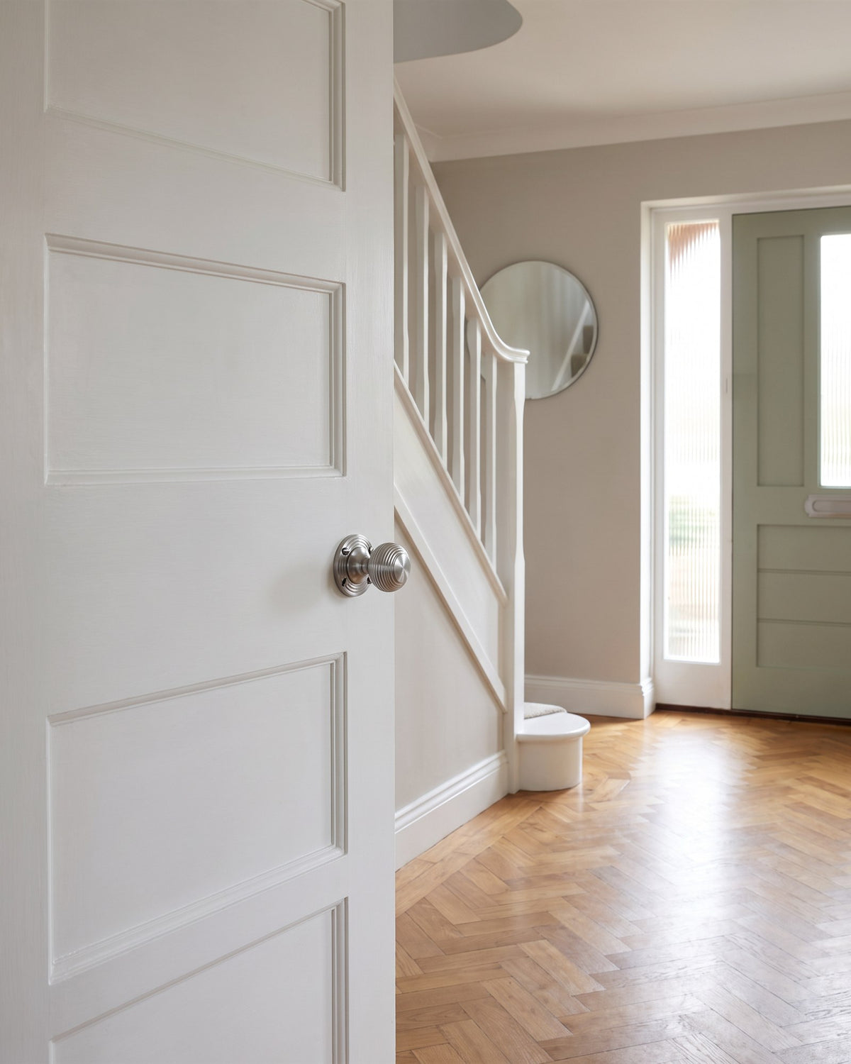 1930s hallway with herringbone parquet and a curved staircase, a satin chrome reeded knob on a white panelled door and the sage front door beyond