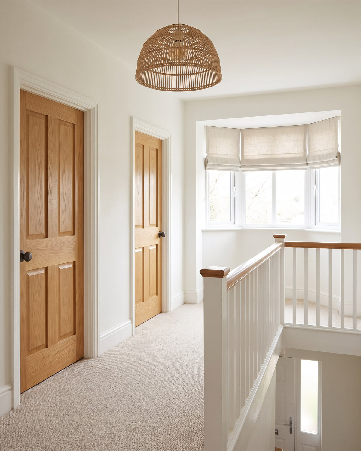 1930s landing with oatmeal wool carpet, a white banister with oak handrail, a rattan pendant and golden oak doors with matt bronze knobs