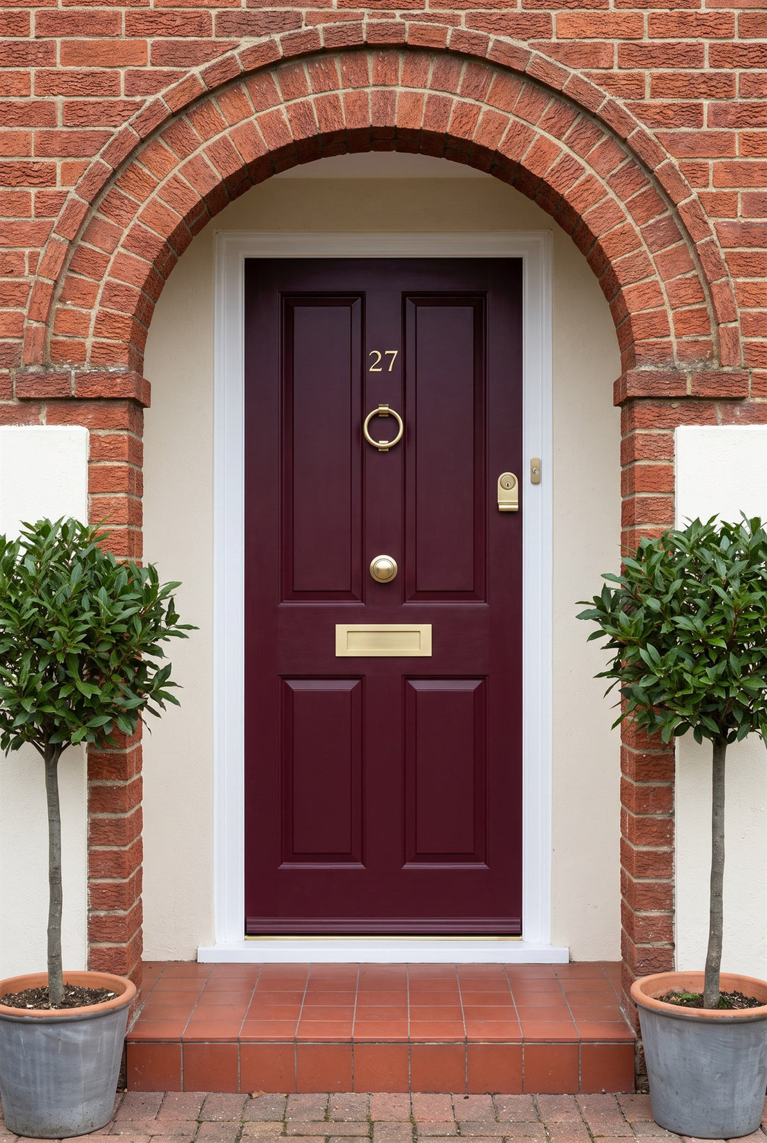Burgundy front door in a 1930s brick porch with bay trees, fitted with satin brass numbers, ring knocker, centre knob, letter plate and cylinder pull