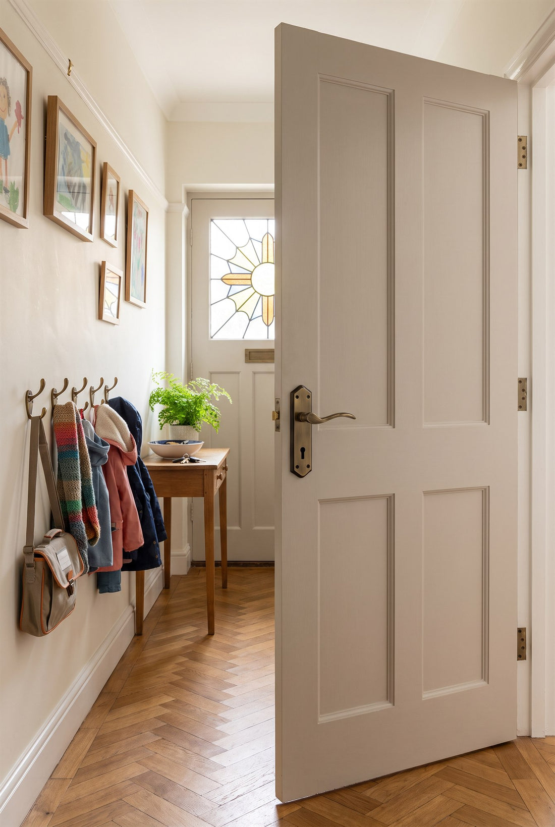1930s hallway with herringbone parquet and a sunburst front door, a greige panelled door with a Paris antique brass and bronze lever lock handle and antique brass hinges, and coats on antique brass hat and coat hooks