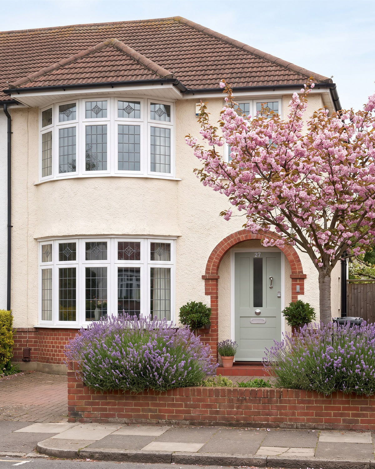 1930s semi with cream pebbledash, a curved leaded bay and a round brick porch arch over a sage front door, lavender and a cherry tree in blossom