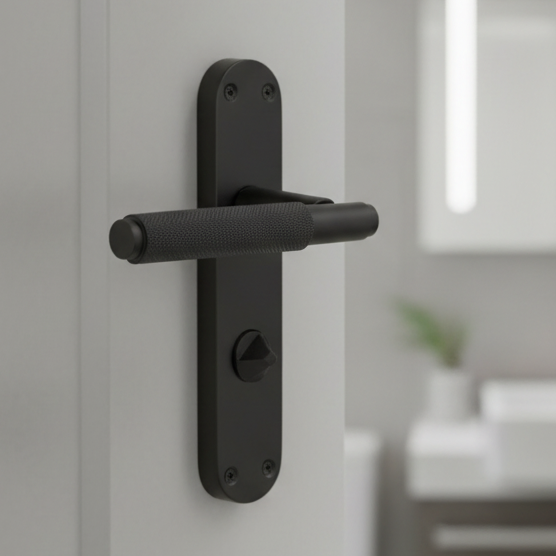 Matt black knurled bathroom handle with a privacy turn on a long rounded backplate, a white bathroom behind