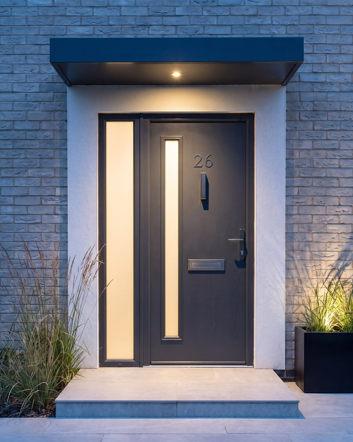 Anthracite composite front door at dusk lit by a canopy downlight, warm light through the frosted glass strip and graphite fittings catching the light