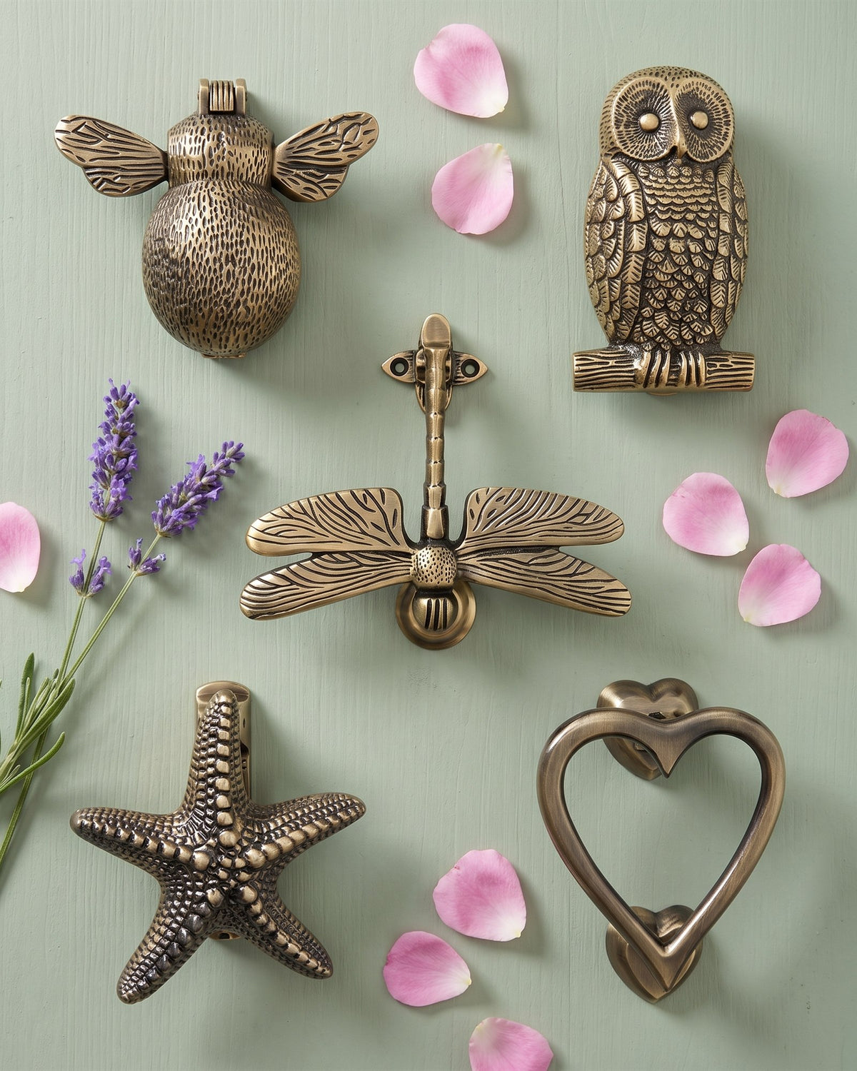 Antique brass bee, owl, dragonfly, starfish and heart door knockers laid out on a sage painted board with lavender and rose petals