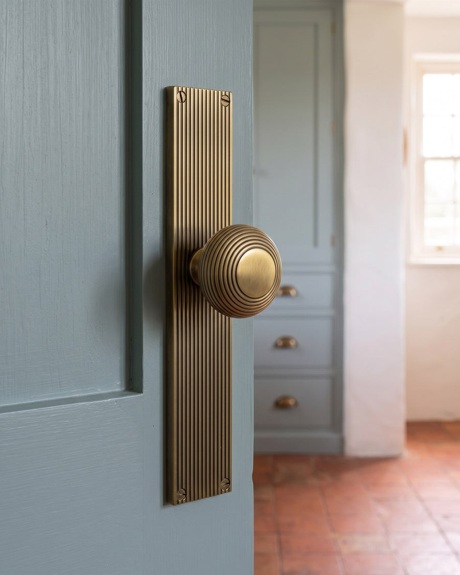 Antique brass beehive knob on a reeded latch plate on a blue-grey green kitchen door, with the larder unit and terracotta floor beyond
