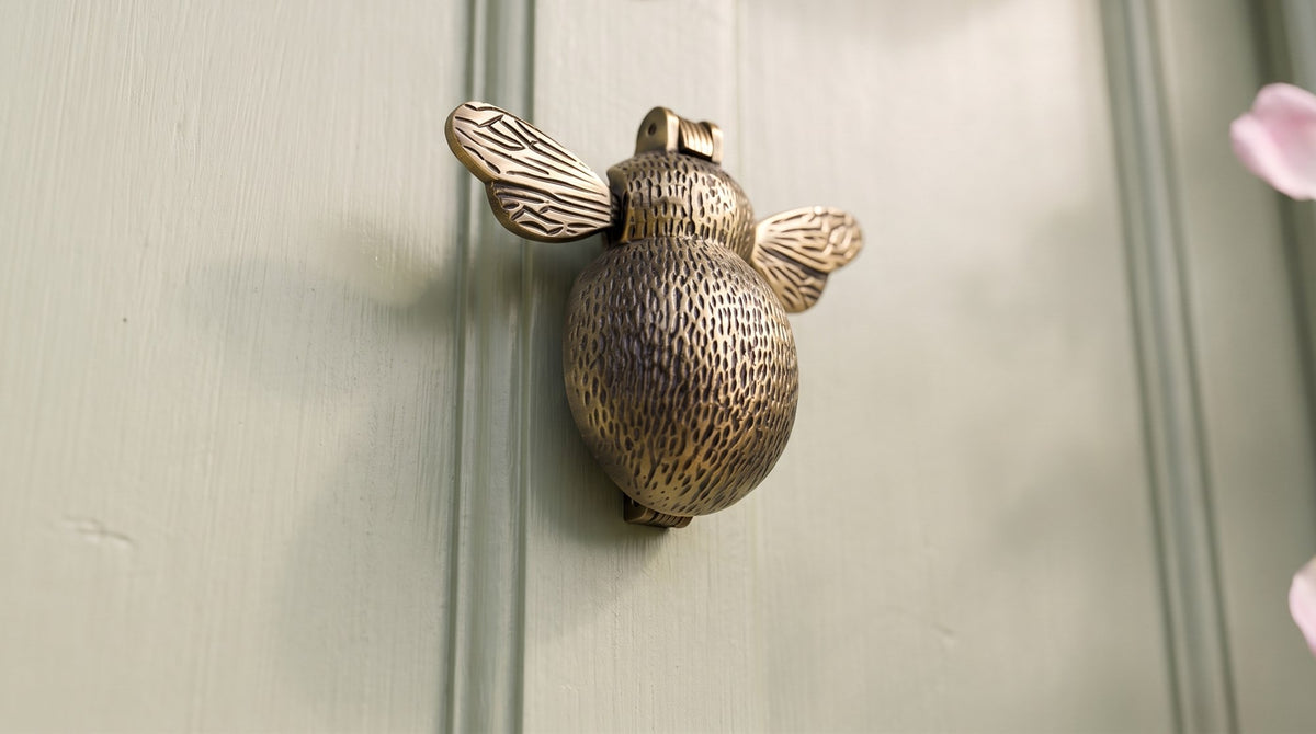 Antique brass bumble bee door knocker with textured body and veined wings on a soft sage green cottage door