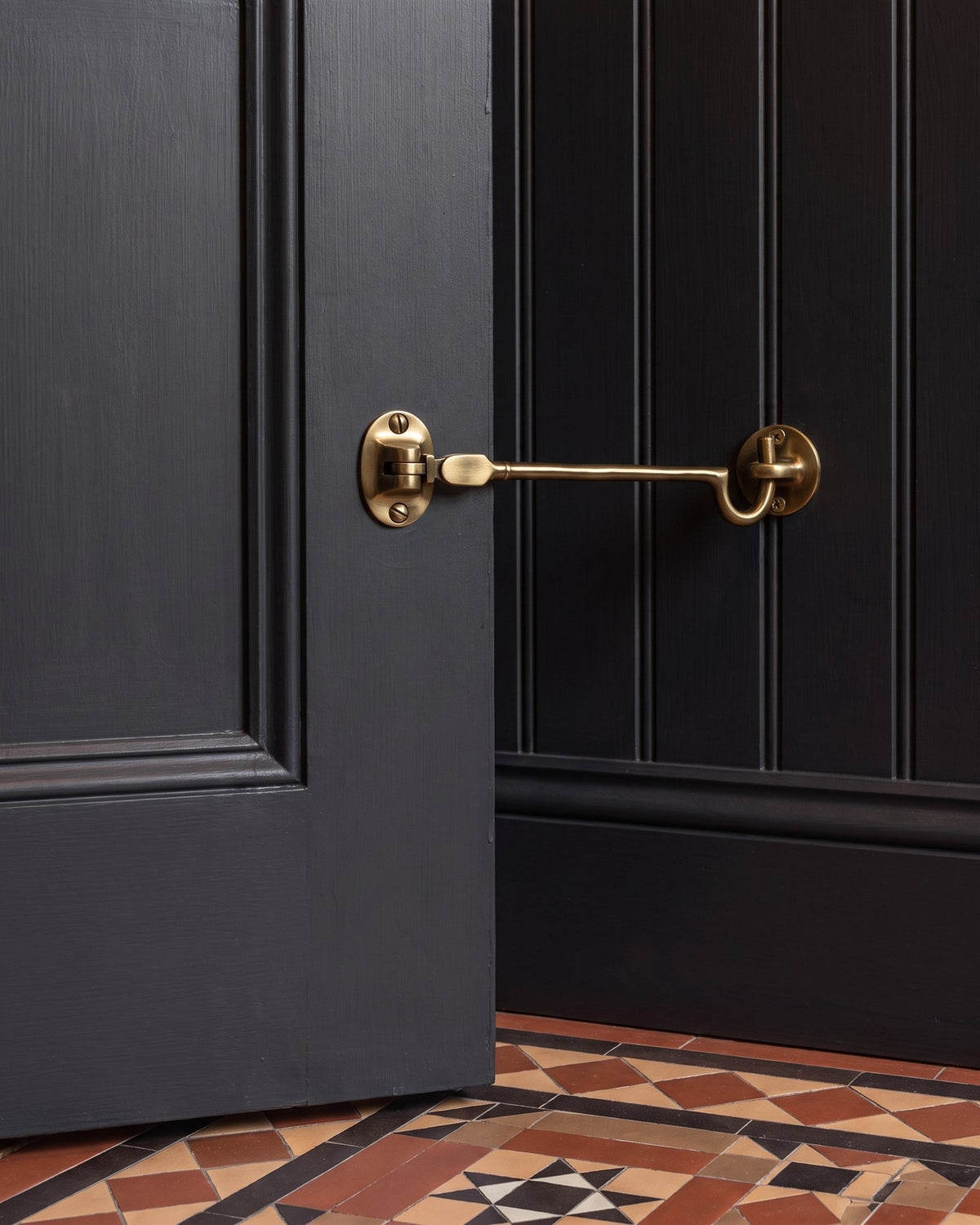 Antique brass cabin hook holding an off-black panelled door open against tongue-and-groove panelling above encaustic tiles