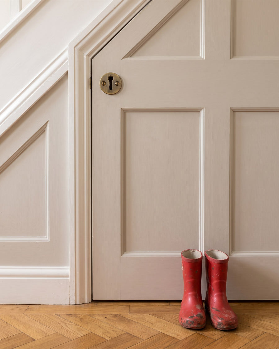 Antique brass circular keyhole escutcheon on a greige panelled understairs cupboard door, red children's wellies below