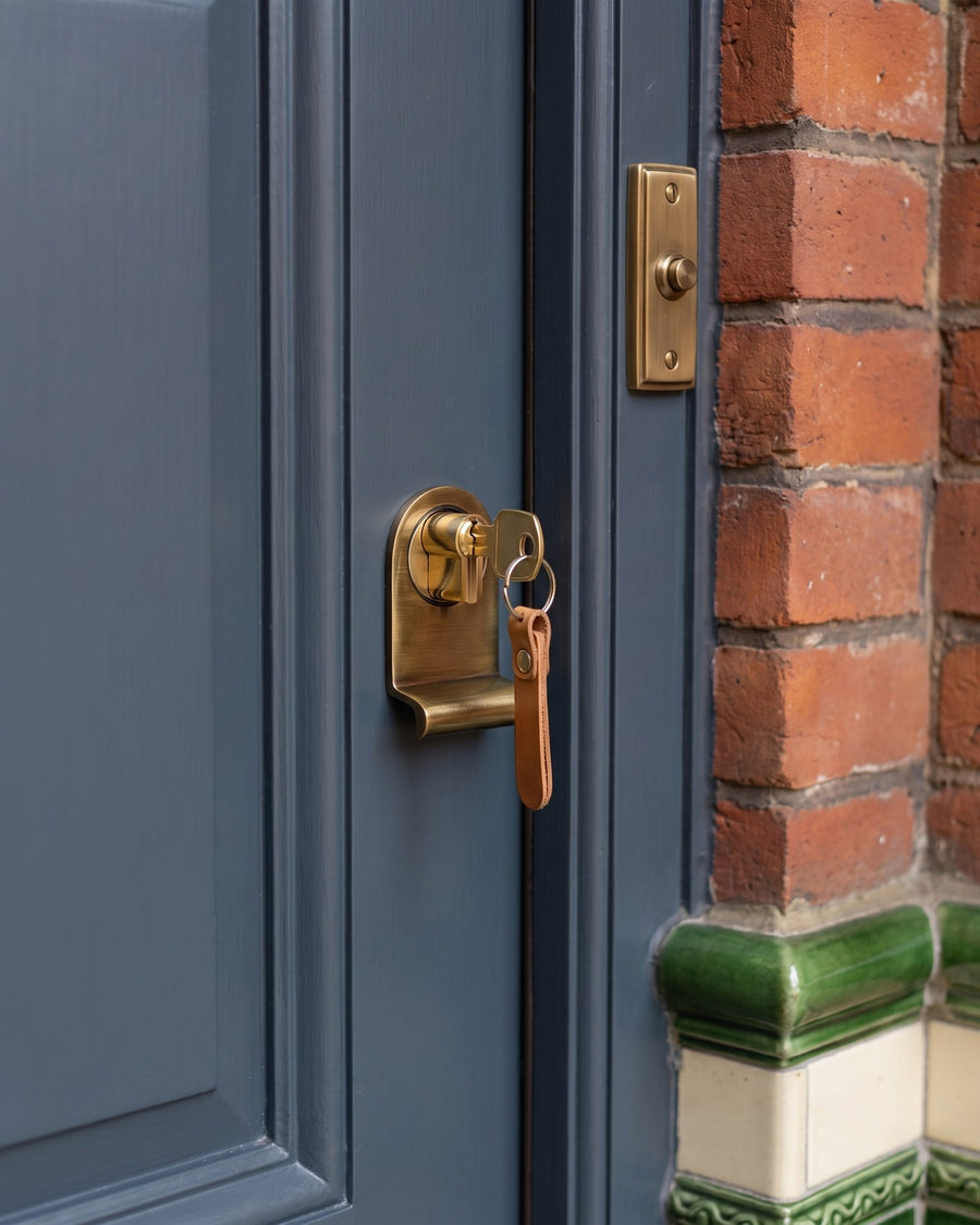 Antique brass cylinder pull with a key on a tan leather fob in the cylinder, beside an antique brass bell push on the brick porch reveal