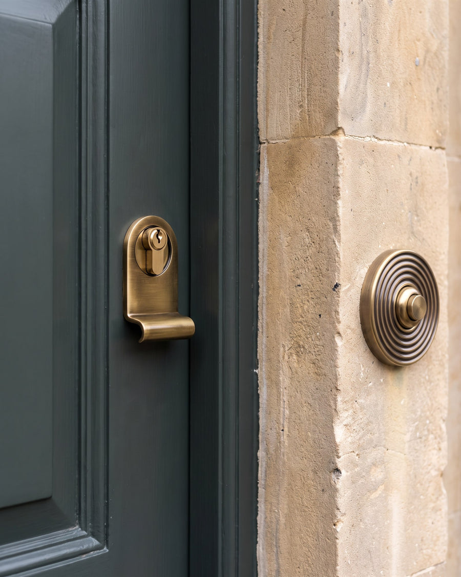Antique brass cylinder pull with a brass euro cylinder on a green door beside a round reeded antique brass bell push on a Bath stone pilaster