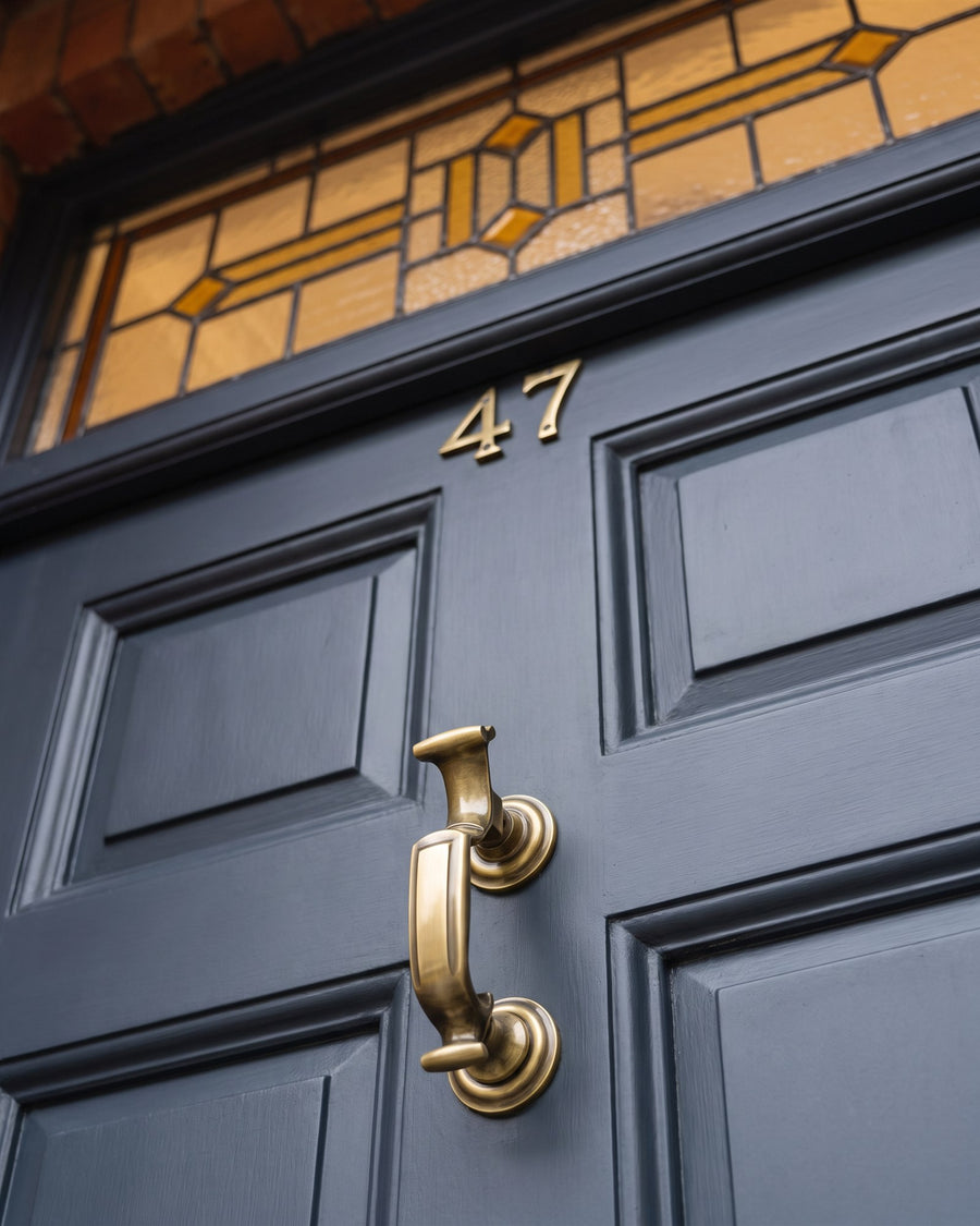 Antique brass doctor knocker and serif numerals 47 on a grey four-panel door beneath an amber leaded stained-glass fanlight