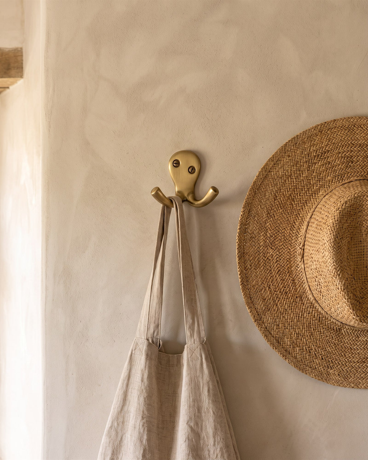 Antique brass double coat hook holding a linen tote on a cloudy putty limewashed wall
