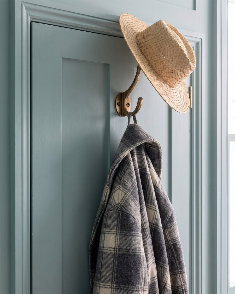 Antique brass hat and coat hook on the back of a blue-green bedroom door holding a straw hat and a checked wool coat