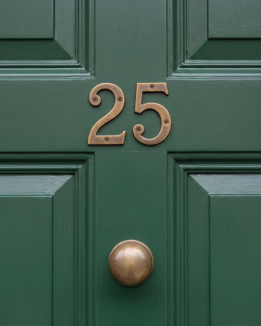 Antique brass serif house numbers 25 on the rail of a racing green panelled front door above an antique brass round knob