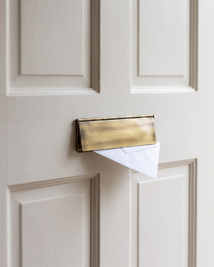 Antique brass interior flap with an envelope beneath it on the inside of an off-white four-panel cottage door