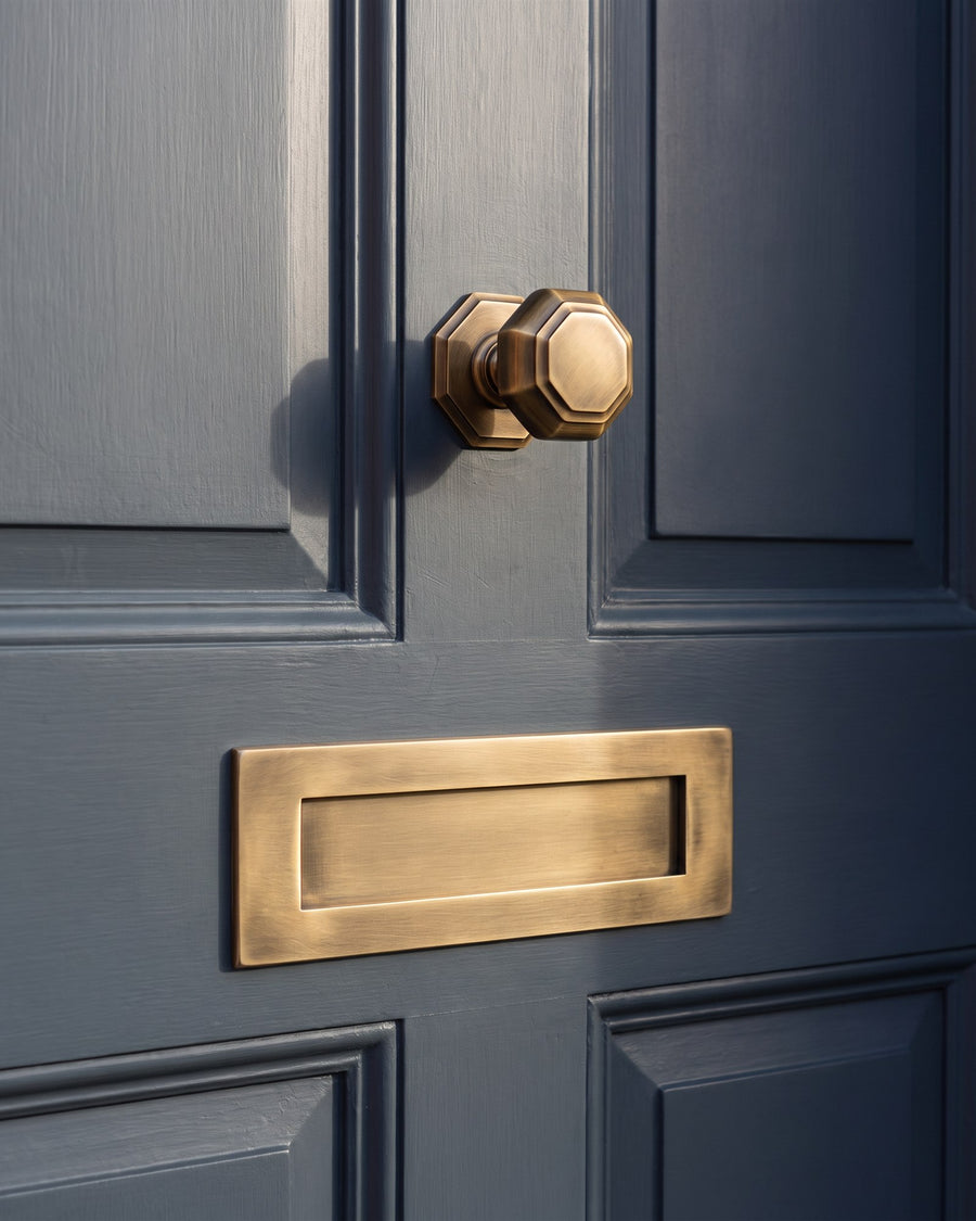 Wide antique brass letter plate on the middle rail of a lead-grey door with the octagonal centre knob above it in low sunlight