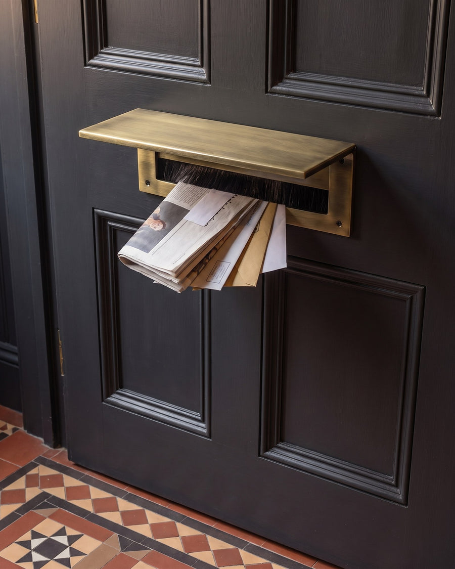 Antique brass internal letter tidy with brush draught excluder on the inside of an off-black front door, with post dropping through