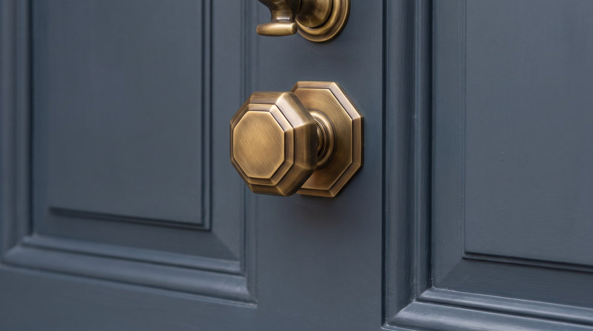Antique brass octagonal centre door knob on its octagonal base, below the end of an antique brass doctor knocker, on a dark blue-grey painted Victorian door