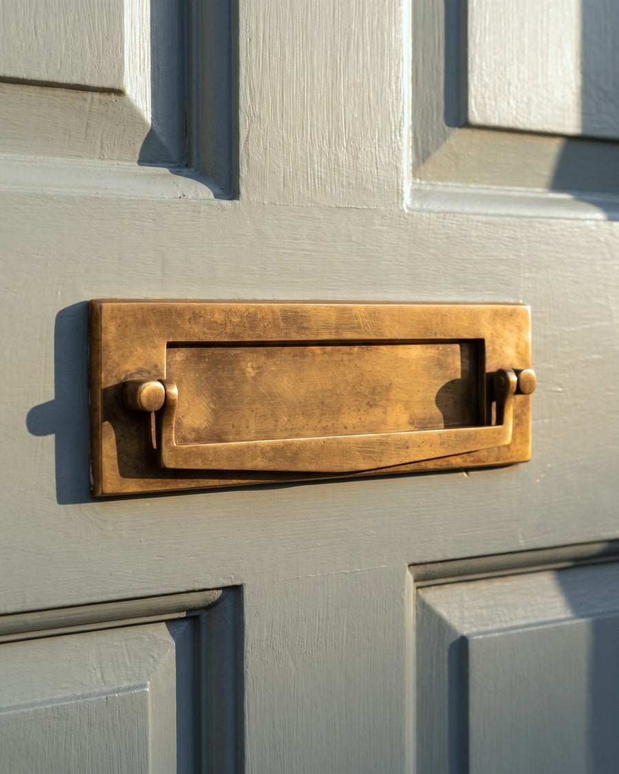 Antique brass postal knocker letter box, whose hinged flap doubles as a door knocker, on a grey-green painted door in low sun
