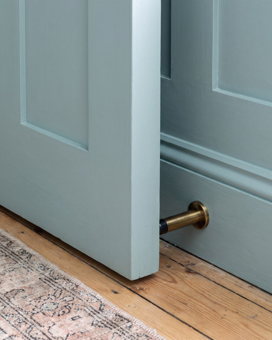 Blue-green panelled door resting against an antique brass projection door stop on a moulded skirting above pine floorboards