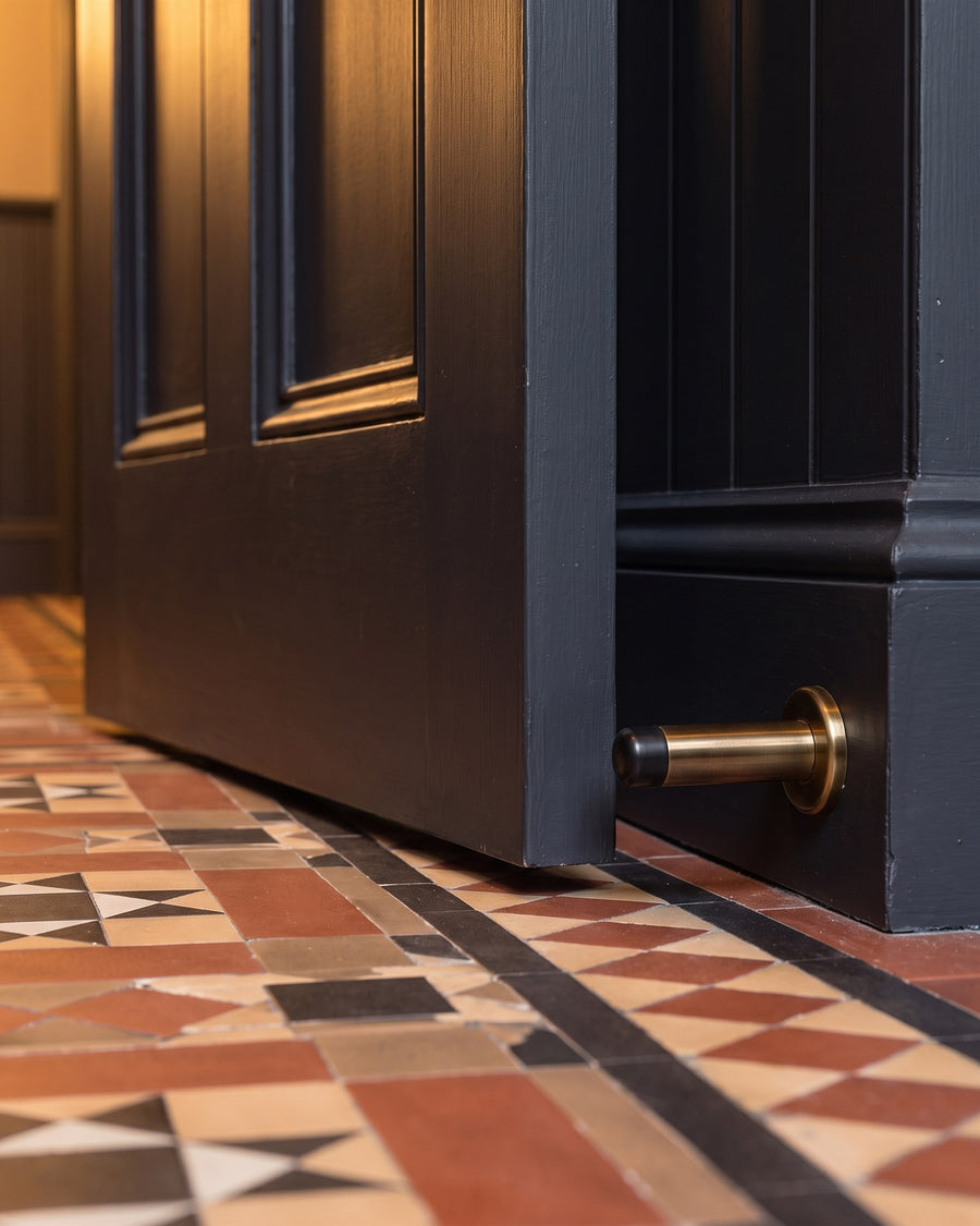 Off-black panelled door resting against an antique brass projection door stop fixed to the skirting above encaustic tiles
