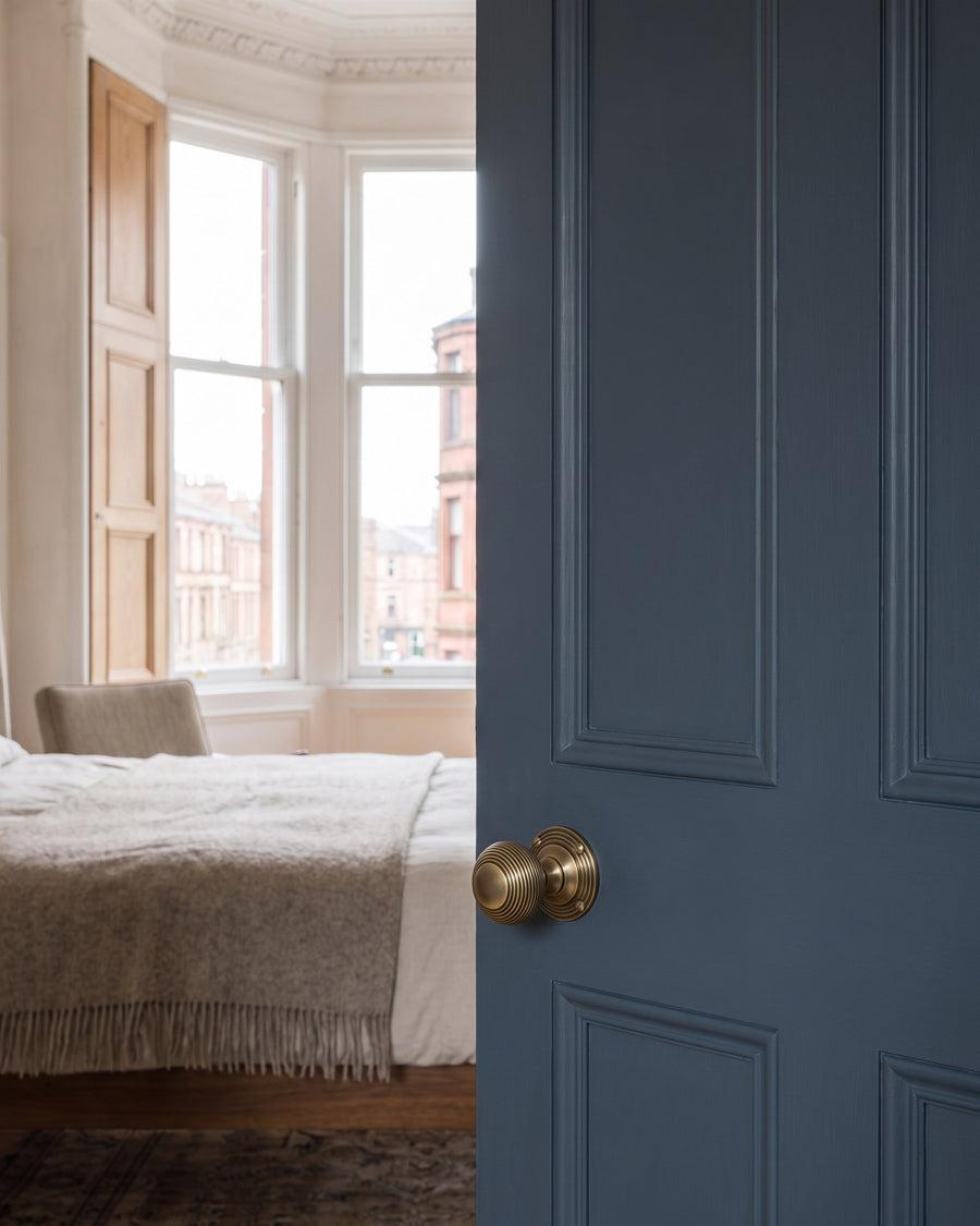 Antique brass reeded beehive mortice knob on a tall blue-grey bedroom door, bay window and bed beyond