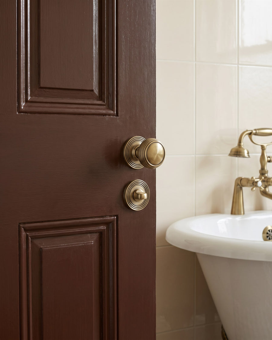 Antique brass reeded knob and reeded turn on a chocolate brown bathroom door beside a roll-top bath and brass tap