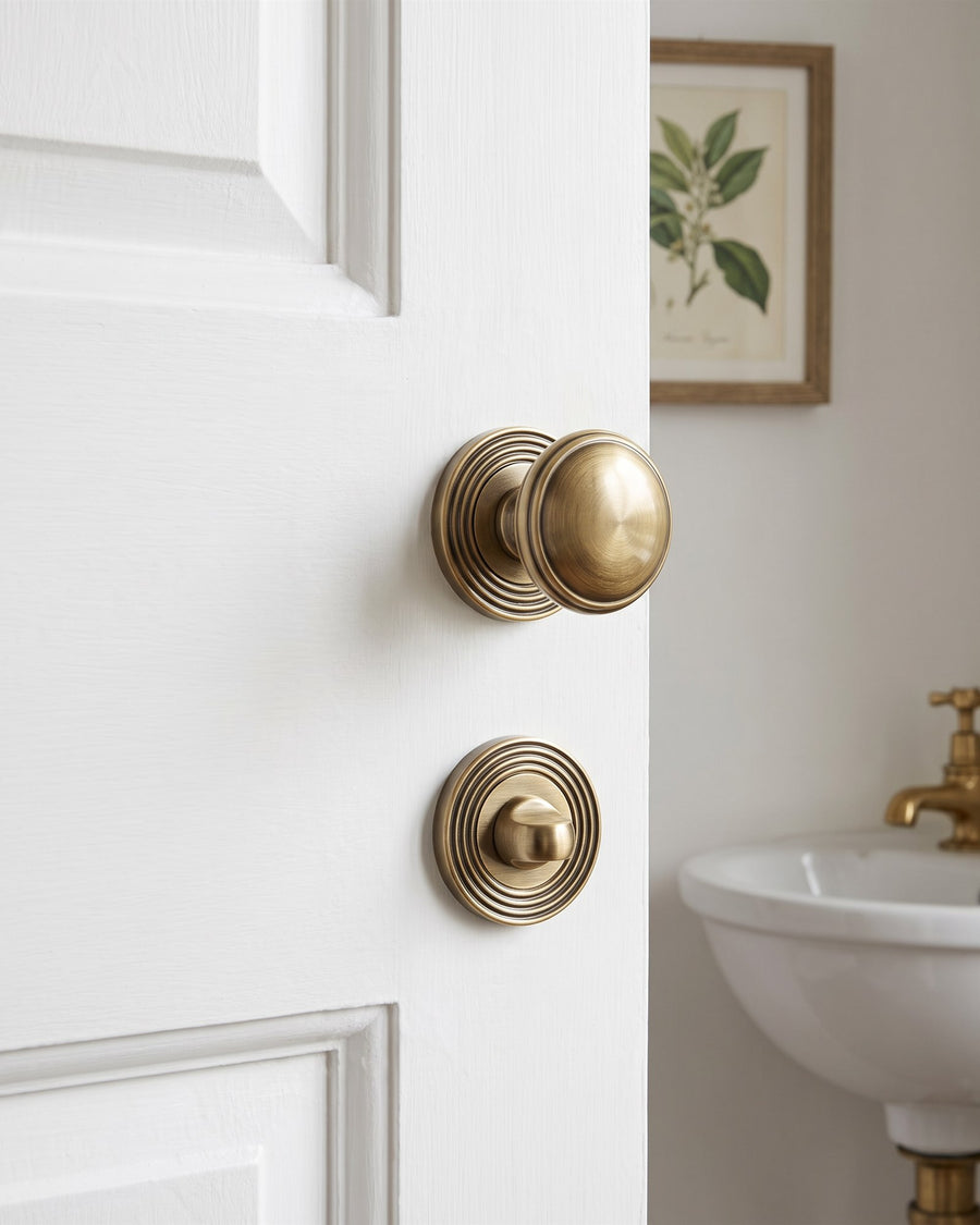 Antique brass reeded mortice knob above a reeded turn and release on a white cloakroom door, a small basin and botanical print beyond