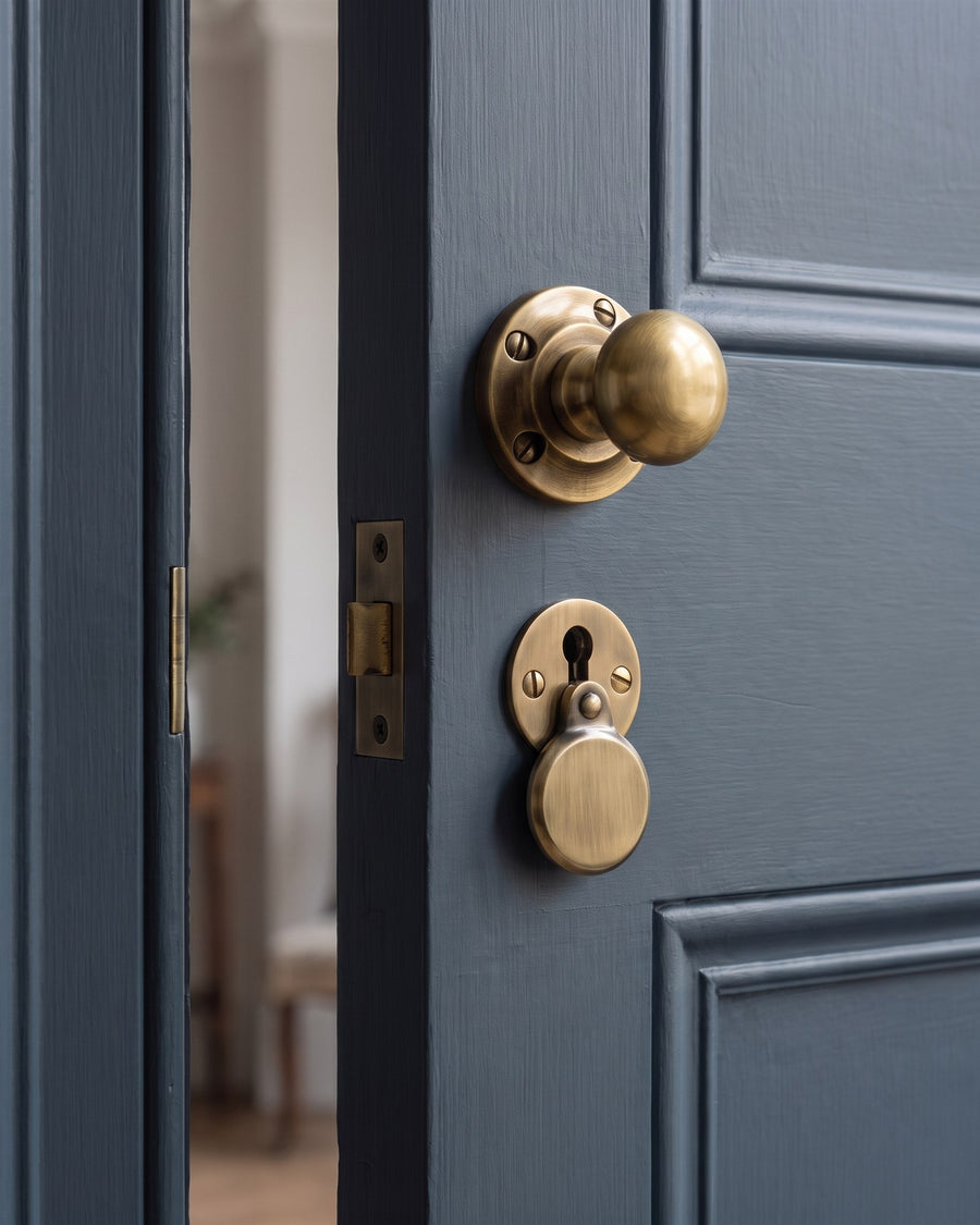 Antique brass Victoria mortice knob with an antique brass heavy covered escutcheon directly below it, near the opening edge of a blue-grey panelled tenement door