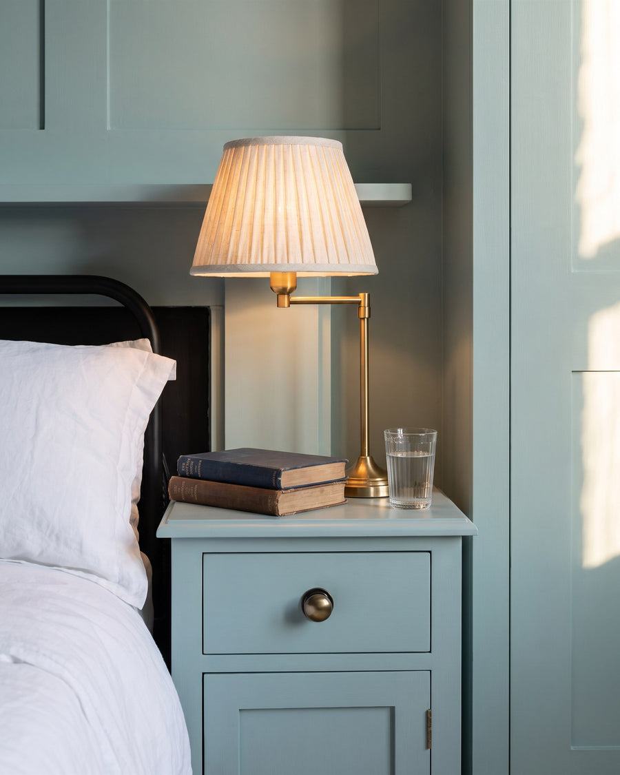 Antique brass domed cabinet knob on a blue-green bedside drawer beside white linen, a pleated lamp, books and a glass of water