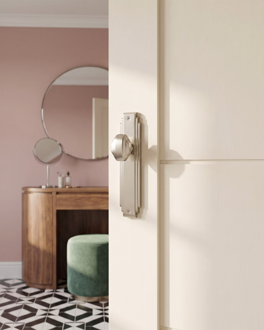 Satin nickel Art Deco knob on a stepped plate on a cream bedroom door, opening onto dusty pink walls, a walnut dressing table and a jade velvet stool