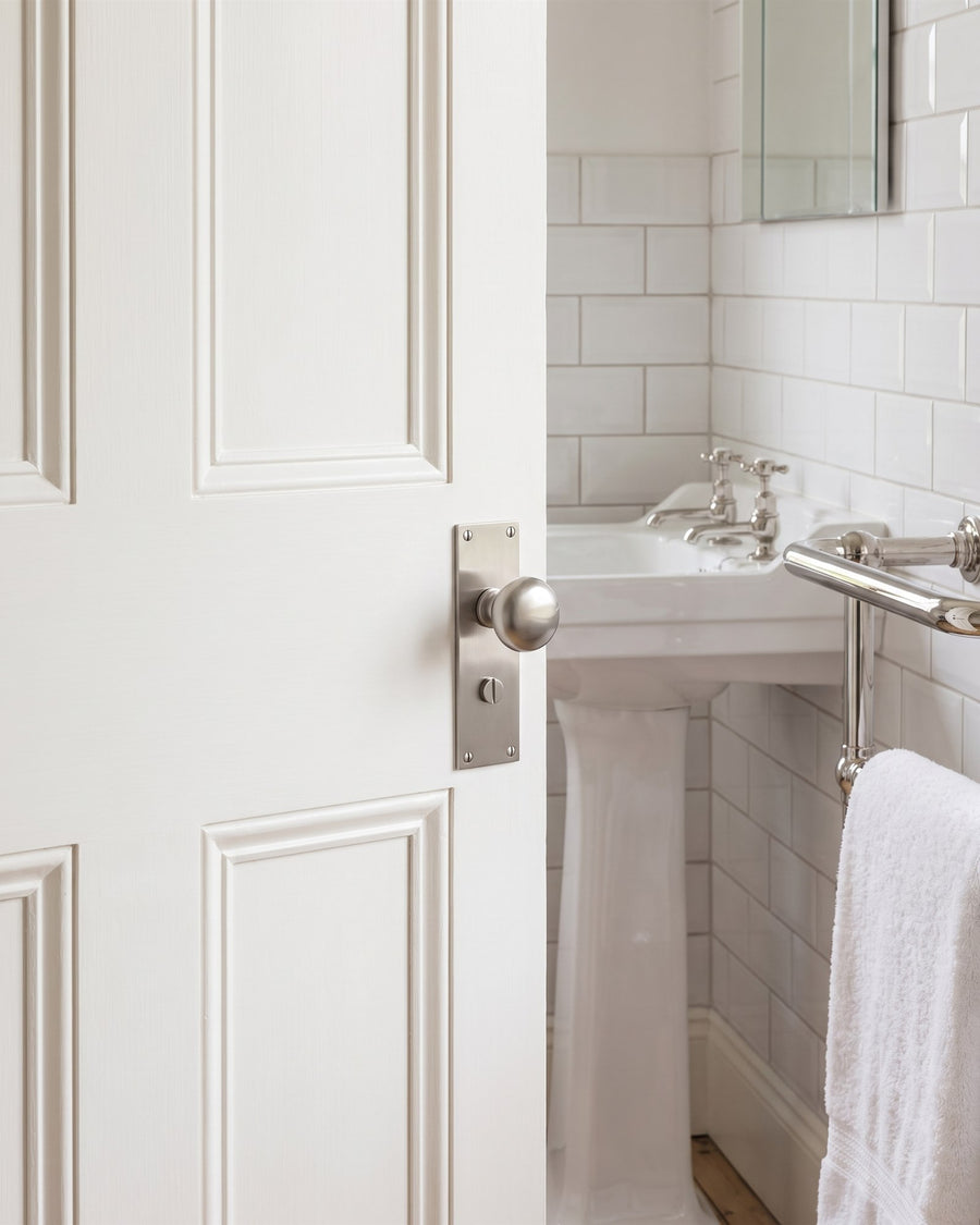 Satin nickel Balmoral knob on bathroom plate with a thumbturn on a white door, white metro tiles, a pedestal basin and nickel towel rail beyond