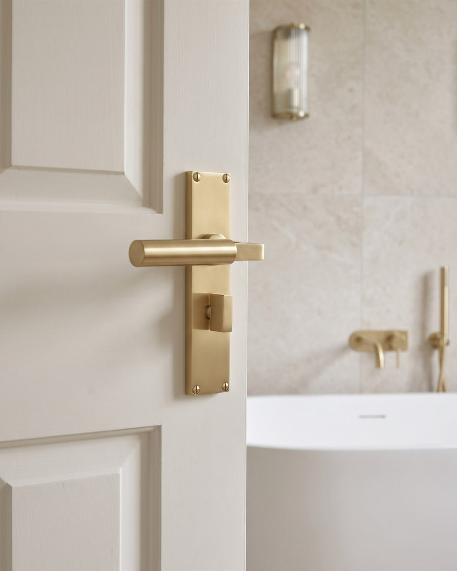 Satin brass Bauhaus bathroom handle with thumbturn on a white door, freestanding bath and brass mixer beyond