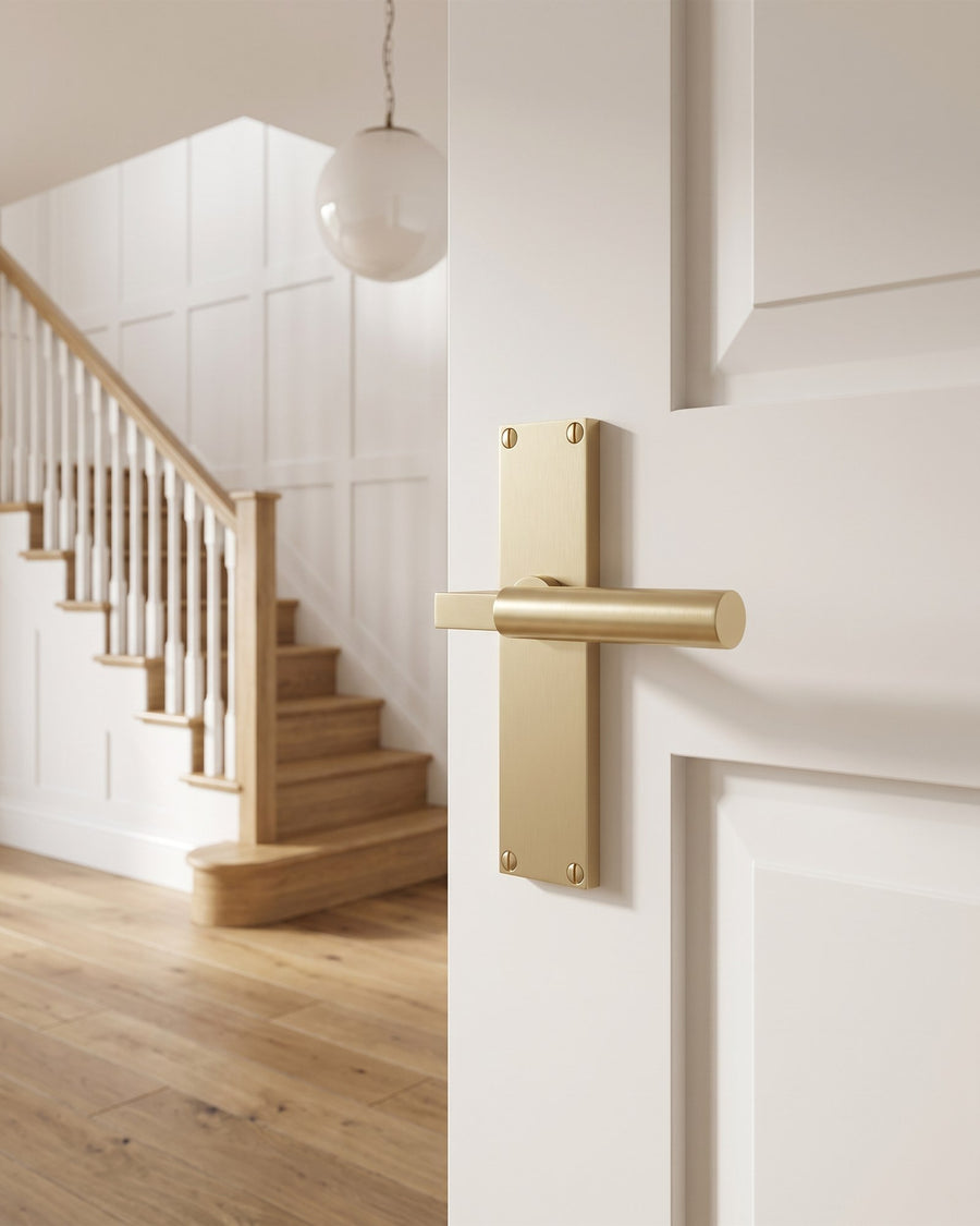 Satin brass Bauhaus lever on a warm white hall door with an oak staircase and globe pendant beyond