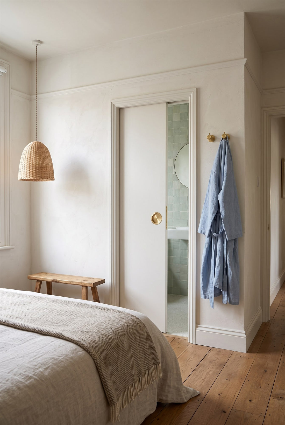 Bedroom in a Victorian flat with a white pocket door sliding open onto a green tiled en suite, fitted with a satin brass circular flush pull and edge pull, and knurled satin brass hooks holding a robe