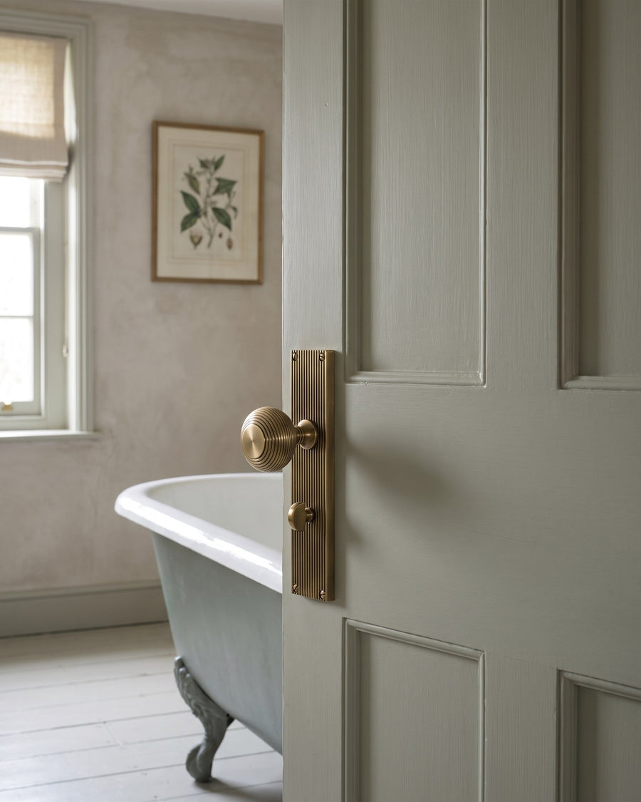 Antique brass beehive knob on a reeded bathroom plate with thumbturn, on a grey-green door opening to a roll-top bath