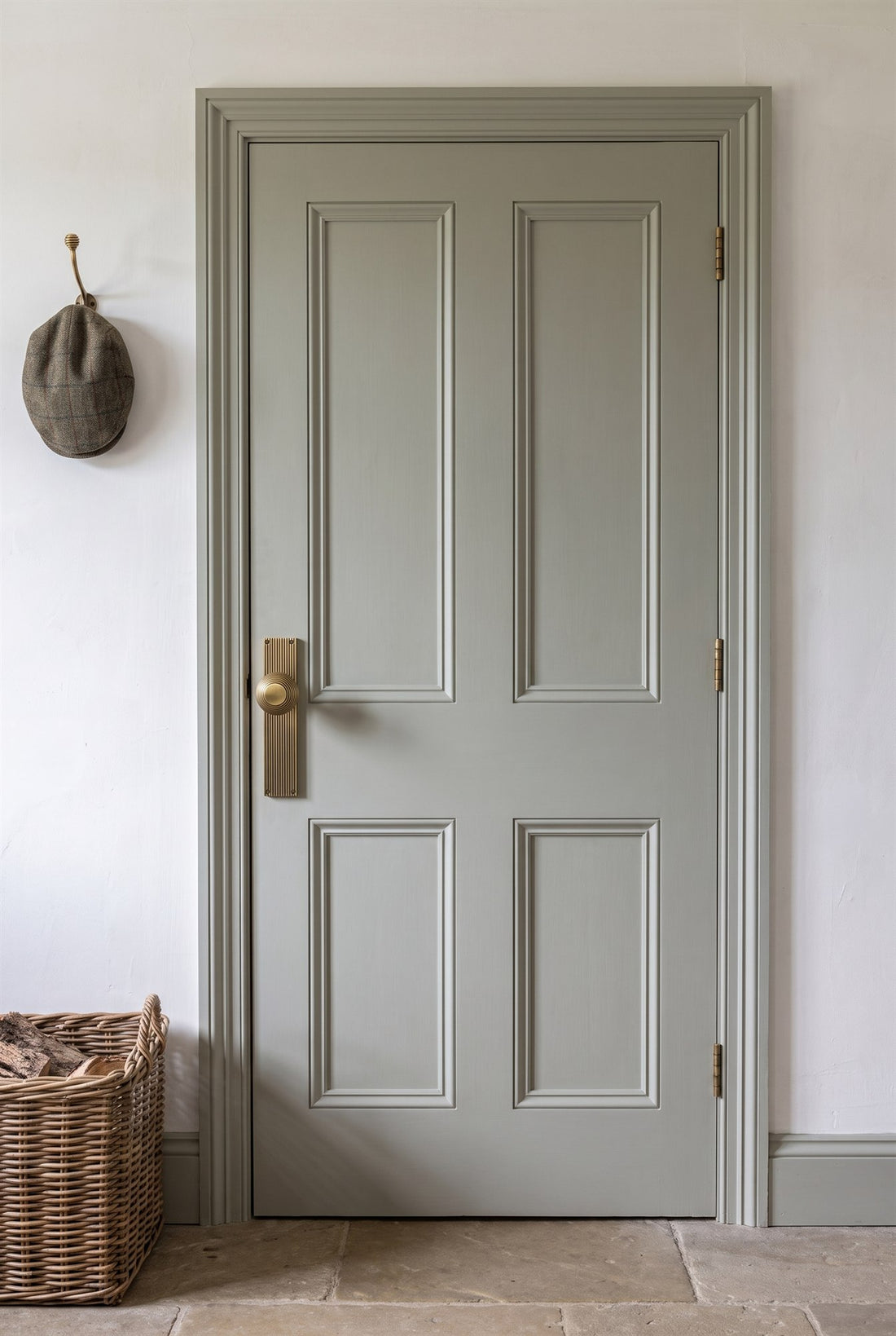 Grey-green four-panel door with an antique brass beehive knob on a reeded plate, antique brass hinges and a beehive coat hook holding a tweed cap on the wall