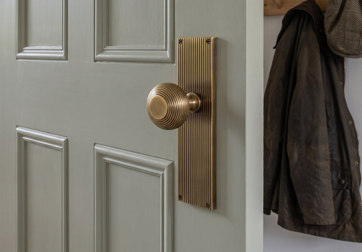 Antique brass beehive knob on a reeded backplate on a soft grey-green panelled door, with a log basket on flagstones and a waxed jacket on a peg rail