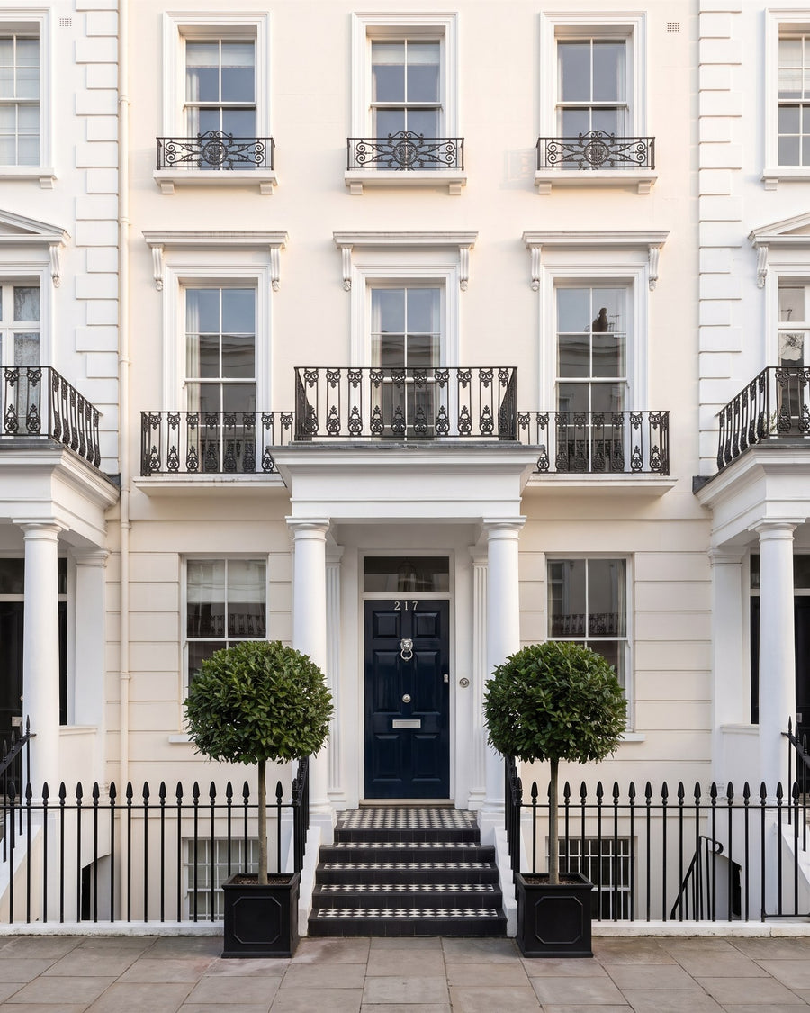 White stucco Regency terrace with balconettes, a columned portico, black railings, chequered steps, bay trees and a navy front door