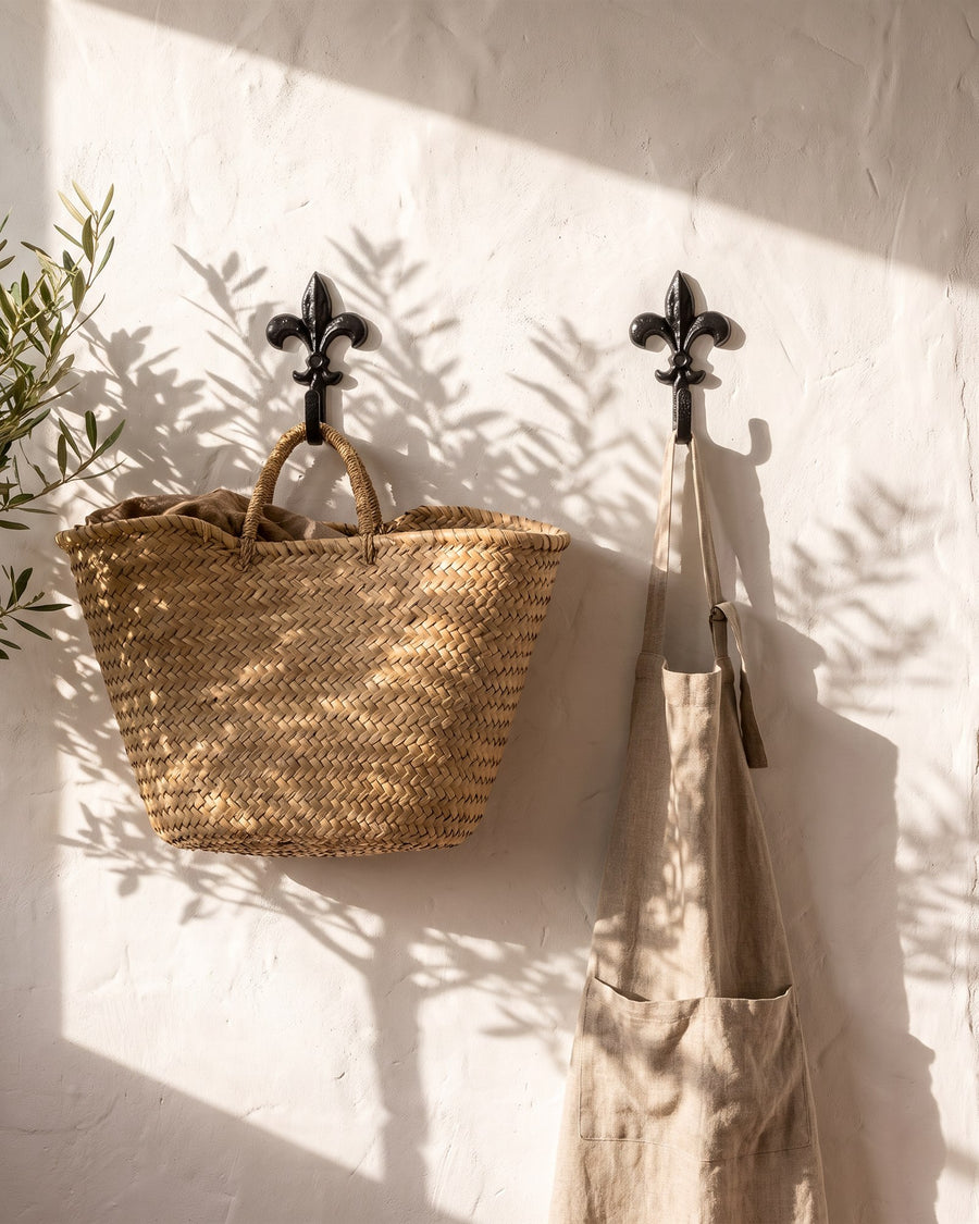Black antique iron fleur-de-lys hooks holding a woven basket and a linen apron on a sunlit limewashed wall