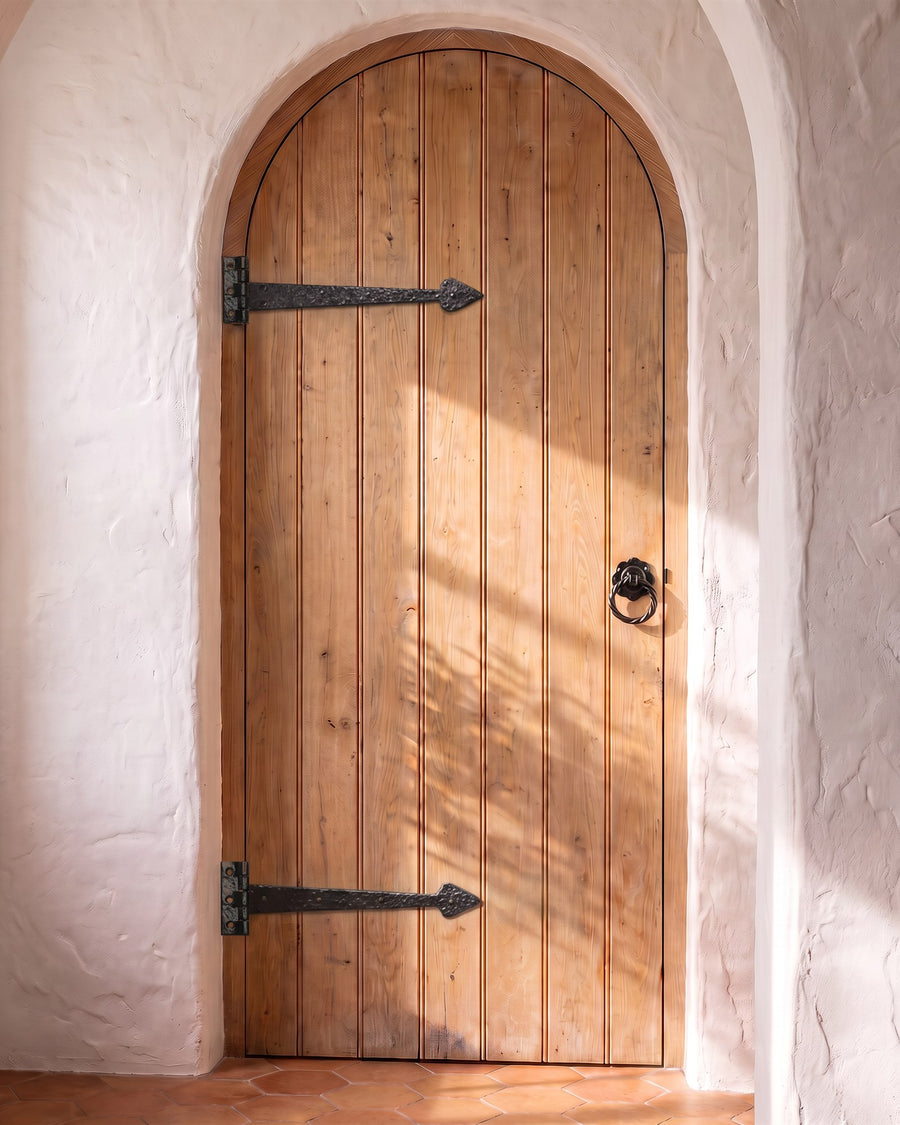 Arched oak plank door in a thick limewashed wall with two black antique iron tee hinges and a black iron twisted ring handle, above terracotta tiles