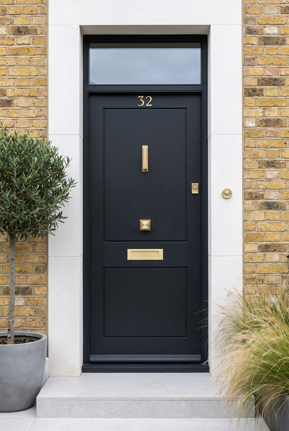 Matt near-black two-panel front door in a white stone surround with satin brass numbers, slim knocker, square centre knob, letter plate, cylinder pull and round bell push