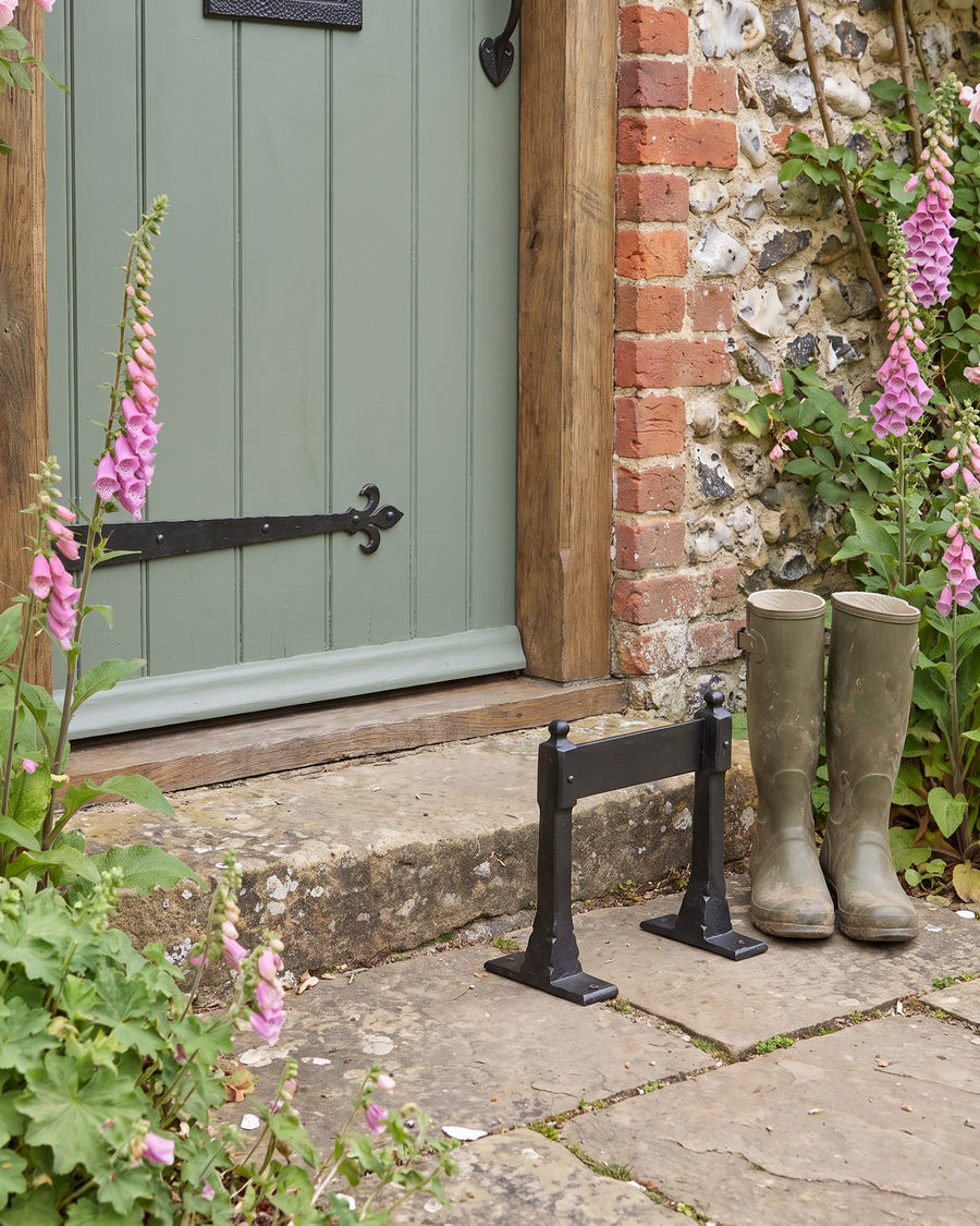 Black iron boot scraper on flagstones beside a worn stone doorstep, with green wellies and foxgloves
