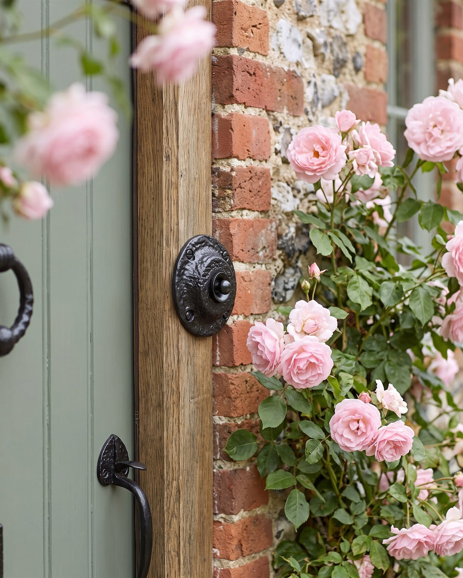 Round black iron bell push on an oak door frame surrounded by pink climbing roses