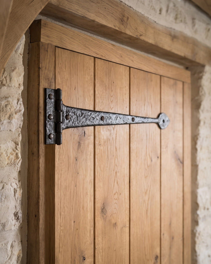 Hammered black iron tee hinge across the planks of an oak ledged door beside a limewashed stone wall