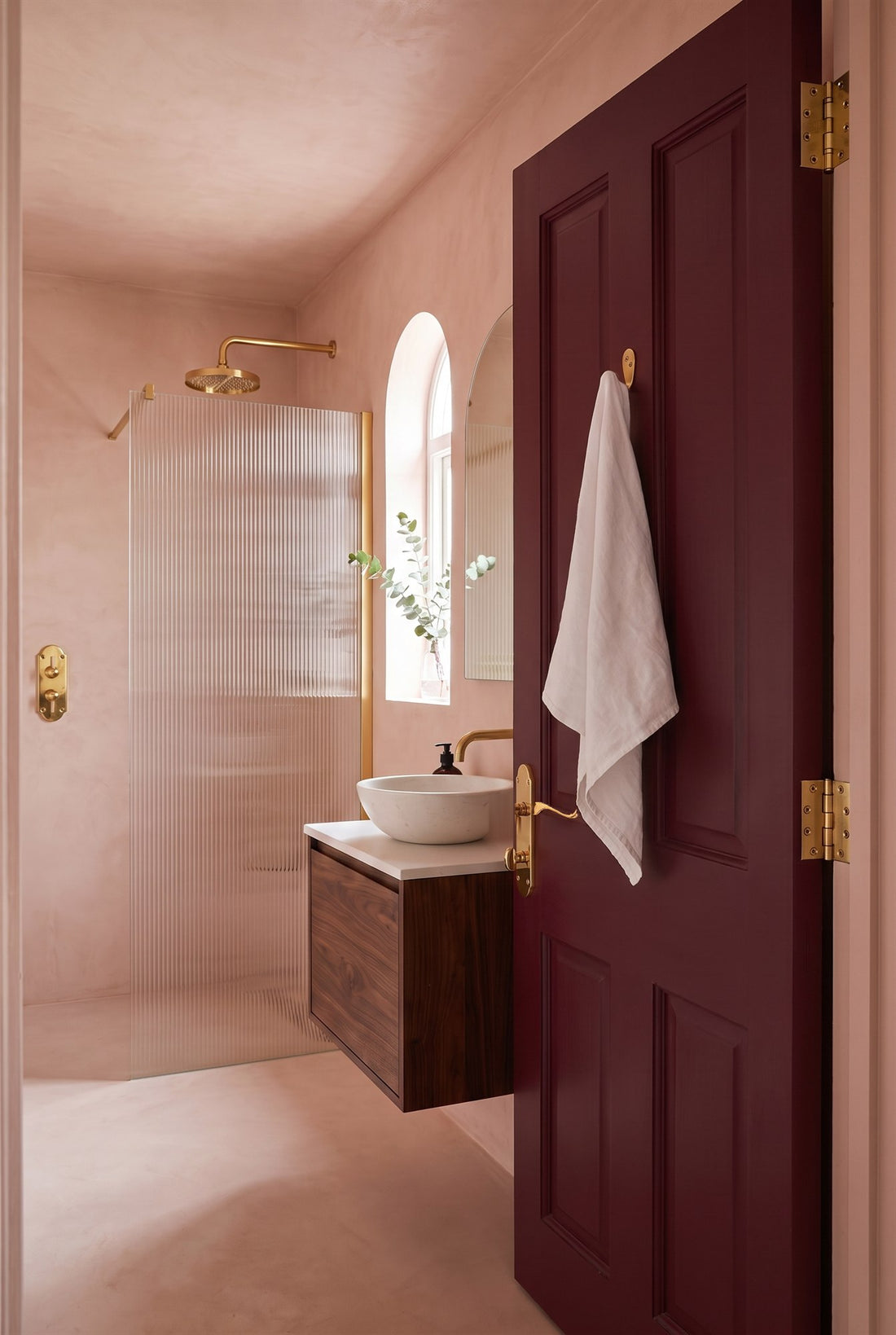Blush micro-cement en suite with a fluted glass shower and walnut vanity, and a burgundy door with a PVD brass scroll bathroom handle, brass ball-bearing hinges and a brass robe hook