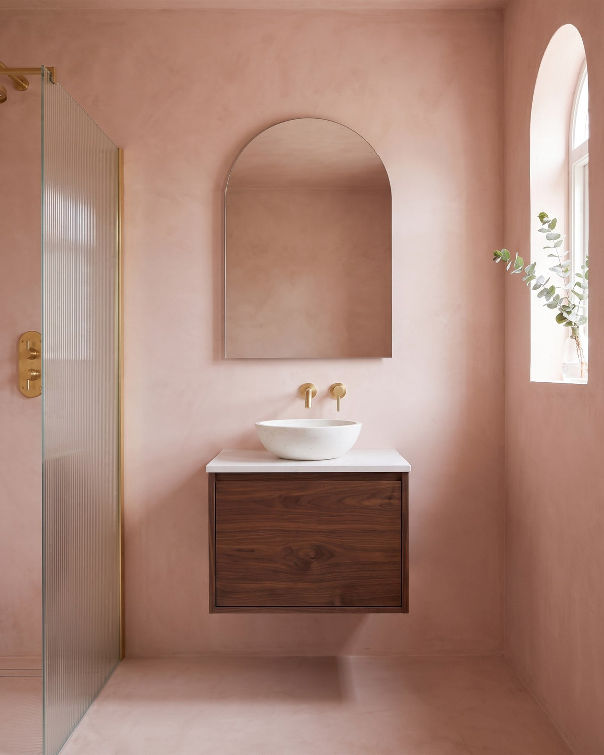 Blush pink micro-cement en suite with a walnut wall-hung vanity, white stone basin, brass tap, arched mirror and fluted glass shower screen